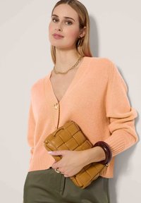 Perzikkleurige v-hals cardigan met een knoopsluiting, gecombineerd met een bruine gevlochten clutch en een chunky armband, tegen een neutrale achtergrond.