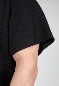 Zwarte textuur met korte mouwen shirt met een geribbeld patroon, voorzien van een ronde halslijn en een ontspannen pasvorm, weergegeven tegen een neutrale achtergrond.