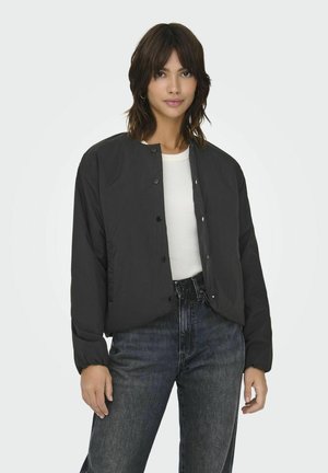 Schwarze Bomberjacke mit rundem Ausschnitt, Druckknöpfen und leicht gepufften Ärmeln. Getragen über einem weißen Hemd und kombiniert mit dunklen Jeans.