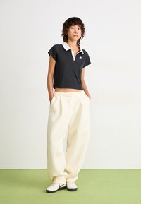 Black cropped polo shirt with white collar, paired with cream wide-leg trousers and white sneakers with black soles. Green floor background.