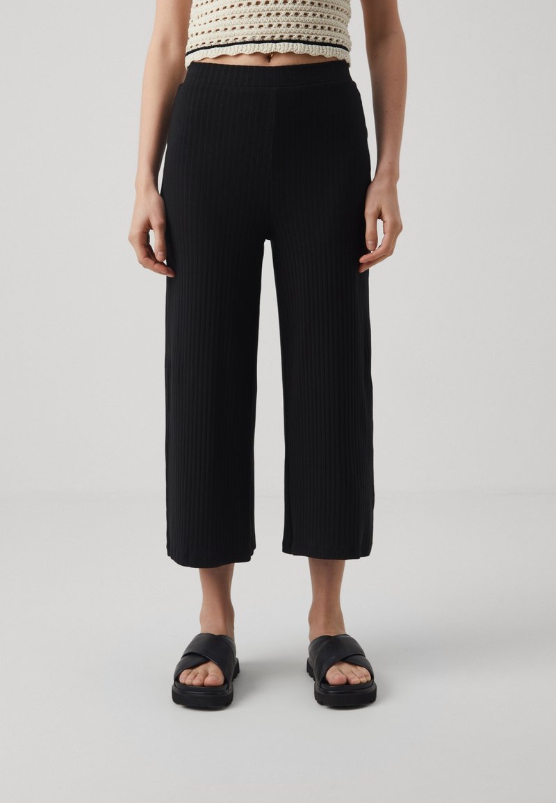Femme portant un pantalon noir large et court, un haut en tricot crème sans manches, et des sandales noires à enfiler sur un fond clair uni.