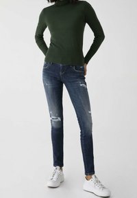 Pull à col roulé côtelé vert associé à un jean skinny déchiré bleu foncé et des baskets blanches, présentant un style décontracté et ajusté.
