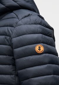 Tengerészkék steppelt textúrájú pehelykabát, kerek narancssárga folttal az ujj felső részén, amelyen "SAVE THE DUCK" felirat szerepel.