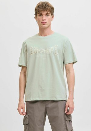 Svijetlozeleni pamučni T-shirt s kratkim rukavima, s natpisom "CORE DEPT." u kontrastnom dizajnu. Uparen s sivim cargo hlačama.