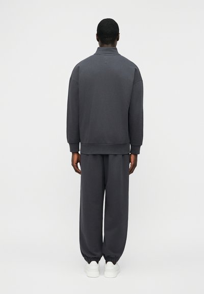 Homme de dos portant un sweat-shirt gris foncé avec manches retroussées et un pantalon de survêtement assorti aux chevilles élastiquées, ainsi que des baskets blanches, sur fond blanc.