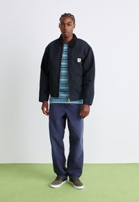 Mann steht auf grünem Boden und trägt eine schwarze Zip-Up-Jacke, ein blau gestreiftes Hemd, dunkelblaue Hosen und grüne Turnschuhe vor einem weißen Wandhintergrund.