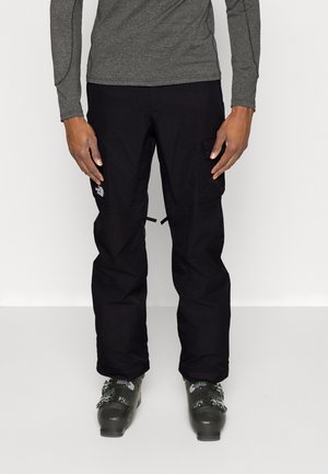 Person trägt schwarze Schneehose mit Cargotaschen und graue Skistiefel, steht vor einem einfarbigen hellen Hintergrund.