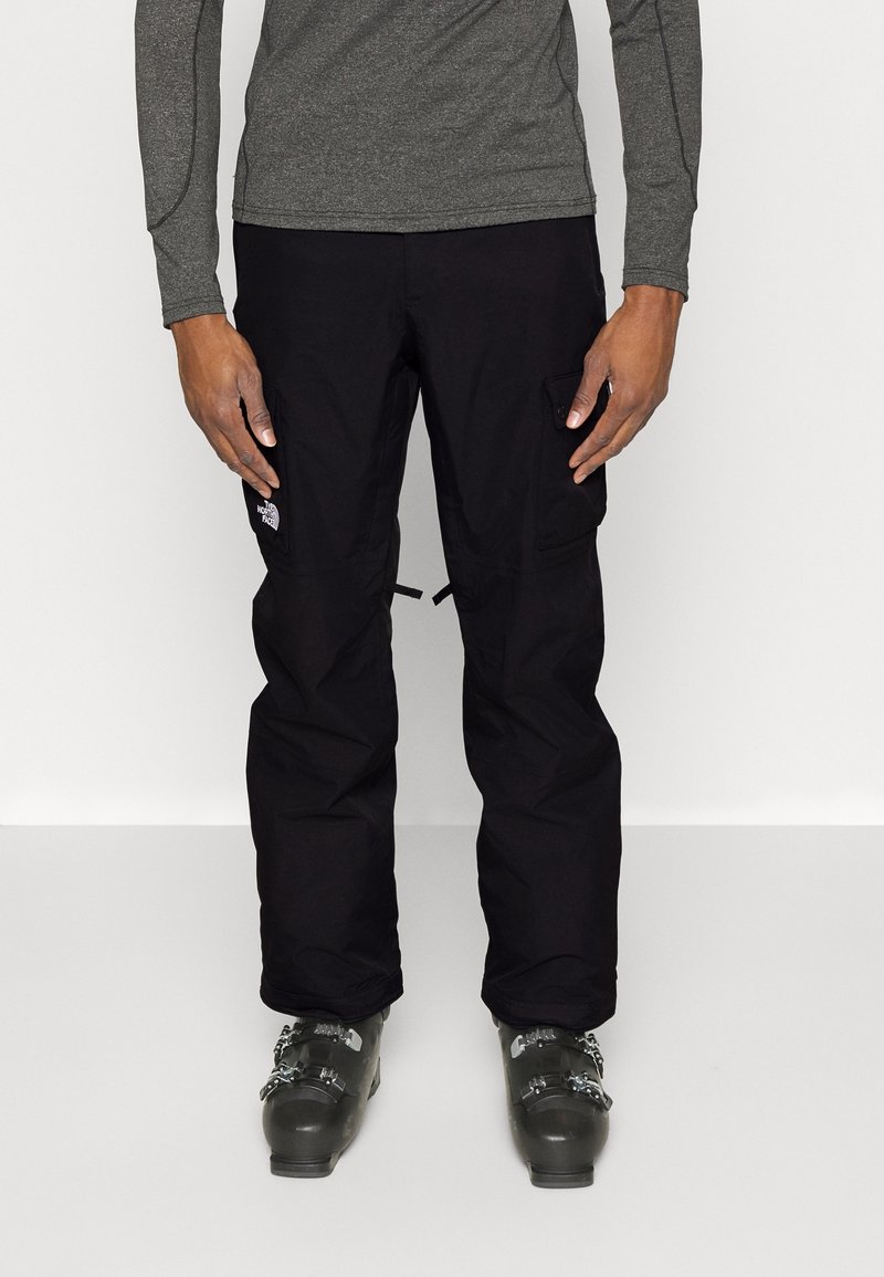 Personne portant un pantalon de neige noir avec des poches cargo et des chaussures de ski grises, debout devant un fond clair uni.
