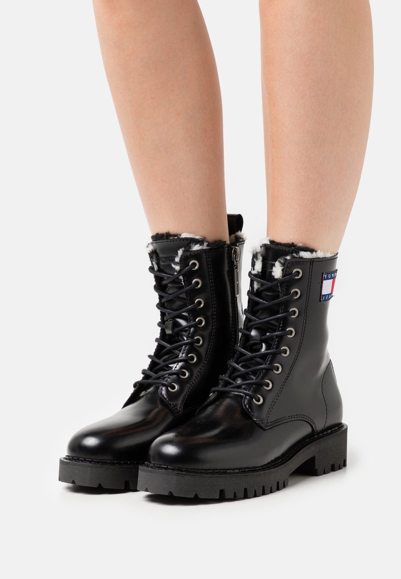 Bottes à lacets en cuir noir avec doublure en fourrure, semelle robuste et fermeture éclair latérale. Comprennent un écusson logo sur la cheville. Texture lisse et design solide.