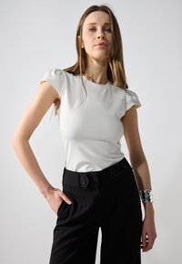 Hvit tettsittende topp med korte puffede ermer, sammen med svarte høyt midje bukser med knappdetaljer og et sølvfarget mansjettarmbånd.