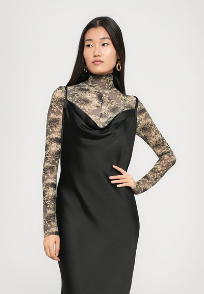 Vrouw met lang, donker haar draagt gouden oorbellen, een doorzichtige bloemenkraagtrui en een zwarte satijnen slipdress, poseert met haar hand op de heup.