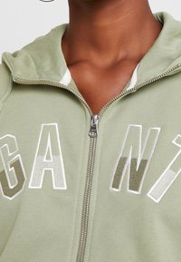 Sudadera con capucha verde con cremallera y texto "AGAIN" bordado en blanco y plateado metálico. Cuenta con una capucha acogedora y tejido texturizado.