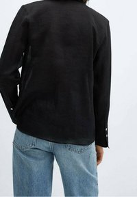 Camisa de lino negra con mangas largas, puños abotonados y dobladillo redondeado, llevada sobre unos pantalones de mezclilla azul claro. Diseño simple y funcional.