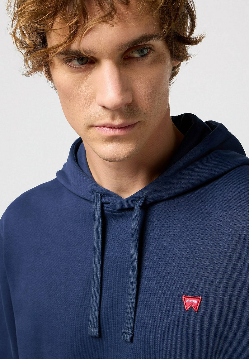 Granatowa bluza z kapturem, wykonana z miękkiego materiału, z sznurkiem w kapturze oraz małym czerwonym logo Wrangler na piersi. Gładka struktura, luźny krój.