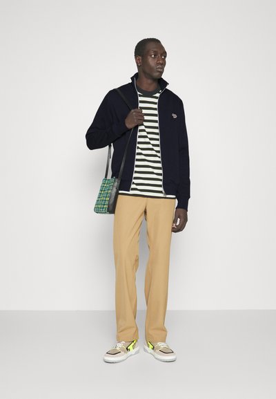 Veste zippée marine, t-shirt rayé, pantalon beige et sneakers multicolores. Tenant un sac à carreaux vert en bandoulière.
