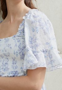 Biała bluzka z kwiecistym wzorem w jasnoniebieskim kolorze, z marszczonym gorsetem, krótkimi falbaniaste rękawami z dodatkowymi falbankami, z miękkiej, teksturowanej tkaniny.