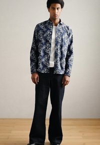 Σκούρο μπλε σακάκι με λευκό λουλουδάτο σχέδιο, φορεμένο πάνω από ένα ελαφρύ πουλόβερ και σκούρα τζιν με φαρδιά μπατζάκια. Λεία υφή, άνετη εφαρμογή.