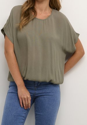 Frau trägt eine locker sitzende olivgrüne kurzärmelige Bluse, die in eine hoch taillierte blaue Jeans gesteckt ist, vor einem einfarbigen Hintergrund.