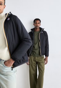 Schwarze Kapuzenjacke mit gelbem Reißverschlussdetail, kombiniert mit einem grünen Hemd und olivfarbigen Hosen. Strukturierter Stoff, leicht gepufftes Design.