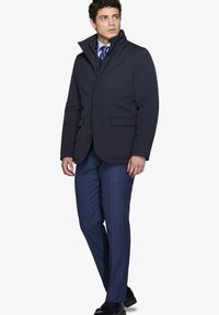 Marinblå isolerad jacka med hög krage och framfickor, i kombination med rutiga blå byxor och svarta skor. Snygg, modern design.