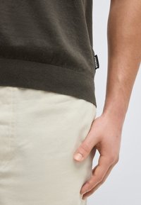 Κοντινό πλάνο του χεριού ενός ατόμου που ακουμπά δίπλα σε παντελόνι κρεμ χρώματος και σκούρο πράσινο πουκάμισο με ετικέτα «ECOALF» στο πλάι.