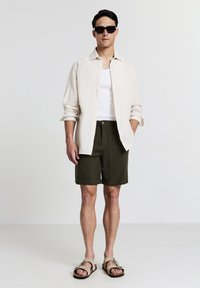 Homme portant un short vert foncé, un débardeur blanc, une chemise beige boutonnée, des sandales beiges et des lunettes de soleil noires, se tenant devant un fond uni.