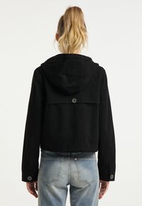 Schwarze cropped Jacke mit Kapuze, knöpfbarem Verschluss und strukturiertem Rückenpartie. Glattes Material mit lockerer Passform, kombiniert mit hellblauen Jeans.