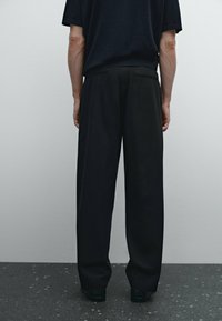 Black wide-leg trousers with a smooth texture, featuring a subtle seam detail at the back and a relaxed fit, paired with a dark shirt.