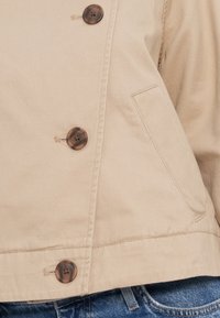 Veste en coton beige avec un design croisé, dotée de quatre boutons sombres et d'une poche latérale. Tissu texturé avec des coutures visibles.