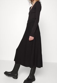 Zwart gebreid langemouwtop met een diepe V-hals, gecombineerd met een vloeiende zwarte midi-rok en robuste zwarte enkelbotjes met textuurpanelen.