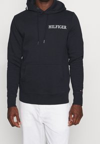 Sudadera azul marino con bolsillo frontal, capucha con cordón y logo blanco "HILFIGER". Presenta puños y dobladillo acanalados, fabricada en tejido suave.