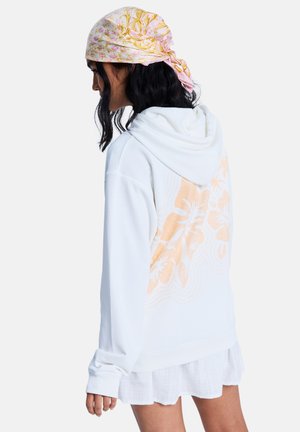 Weißer Hoodie mit großem orangefarbenem Blumenmotiv auf dem Rücken. Mit Kapuze und gerippten Bündchen. Das Modell trägt einen leichten, geschichteten Rock.