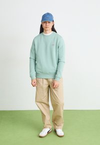 Pullover verde chiaro con polsini a coste, pantaloni khaki e un cappellino in denim blu. Sneakers bianche con dettagli marroni. Design semplice e moderno.
