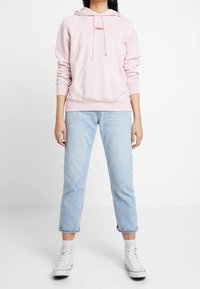Sudadera rosa claro con un bolsillo frontal y capucha con cordón, combinada con jeans azul claro arremangados y zapatillas blancas.