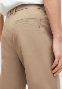 Beige housut, joissa on kapea malli ja jotka on valmistettu teksturoidusta kankaasta. Varustettu takataskulla ja hienovaraisella brodeeratulla kuvioinnilla sivussa.