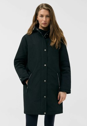 Kobieta z długimi włosami ubrana w czarną, sięgającą kolan parkę z zamkiem błyskawicznym i zatrzaskami, stojąca na tle jednolitego jasnego tła.