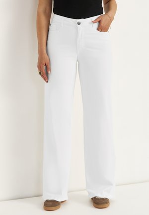 Femme portant un pantalon blanc taille haute à jambes larges avec poches devant, associé à des chaussures décontractées marron et un haut noir.