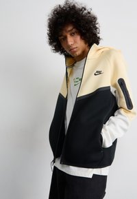 Nike zamšādas jaka ar melnā, krēmkrāsas un baltā krāsā veidotu krāsu bloku. Tekstūrēts audums ar kapuces kabatu ar rāvējslēdzēju un logotipa detaļu.