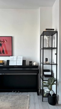 Black Kawai digital piano with sheet music, adjacent black metal shelving unit with decor and potted plant on light wood floor.