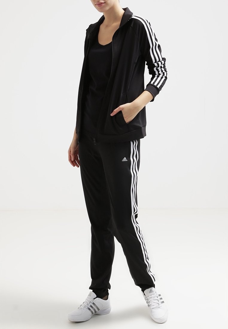 Personne portant un survêtement Adidas noir avec des bandes blanches et des baskets blanches, debout contre un fond uni.