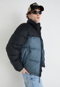 Donsjack met een zwart bovenstuk en een blauw onderstuk, voorzien van een hoge kraag, ritssluiting en zijzakken.