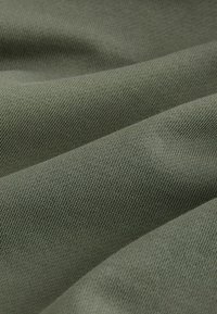 Lähikuva pehmeästä oliivinvihreästä kankaasta, jossa on hieno neulottu rakenne ja lempeitä laskoksia, jotka luovat hienovaraisia varjoja.