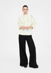 Pullover in maglia chunky di colore crema con colletto alto e motivo in pizzo, abbinato a pantaloni neri a gamba larga, caratterizzati da una texture liscia e fessure laterali.