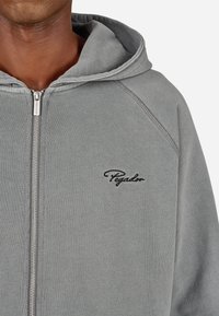 Pelēka kapucīna jakas ar rāvējslēdzēju, izgatavota no mīksta auduma, ar auklu kapuci un izšūtu "Pegador" logo uz krūškurvja. Gluda tekstūra ar smalkām šuvēm.