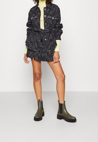 Veste en tweed noir et blanc avec boutons dorés, associée à une jupe noire à volants et des bottines vertes à semelle épaisse.