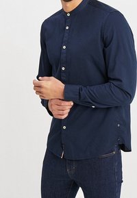 Mann in navyblauer Hemdbluse mit Stehkragen, der das Bündchen zuknöpft, trägt dunkelblaue Jeans und steht vor einem schlichten hellen Hintergrund.