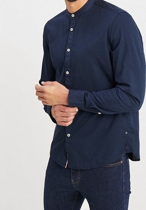 Uomo in camicia blu navy con colletto alla mandarino mentre allaccia il polsino, indossando jeans blu scuro, in piedi contro uno sfondo chiaro e semplice.