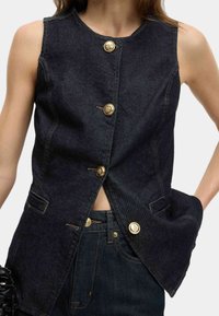 Dunkelblause Jeansweste mit runder Kragenform, ausgestattet mit goldenen Knöpfen, aufgesetzten Taschen und einem Schlitzdetail an der Vorderseite. Glatte Textur.