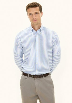 Mężczyzna ubrany w jasnoniebieską i białą koszulę z pionowymi paskami zapinaną na guziki, szare spodnie i ciemny pasek, stojący z rękami za plecami.