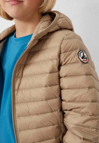 Beige donzen jas met ritssluiting en capuchon, met een doorgestikte textuur. Voorzien van een ronde logopatch op de mouw. Draagt over een blauw overhemd.